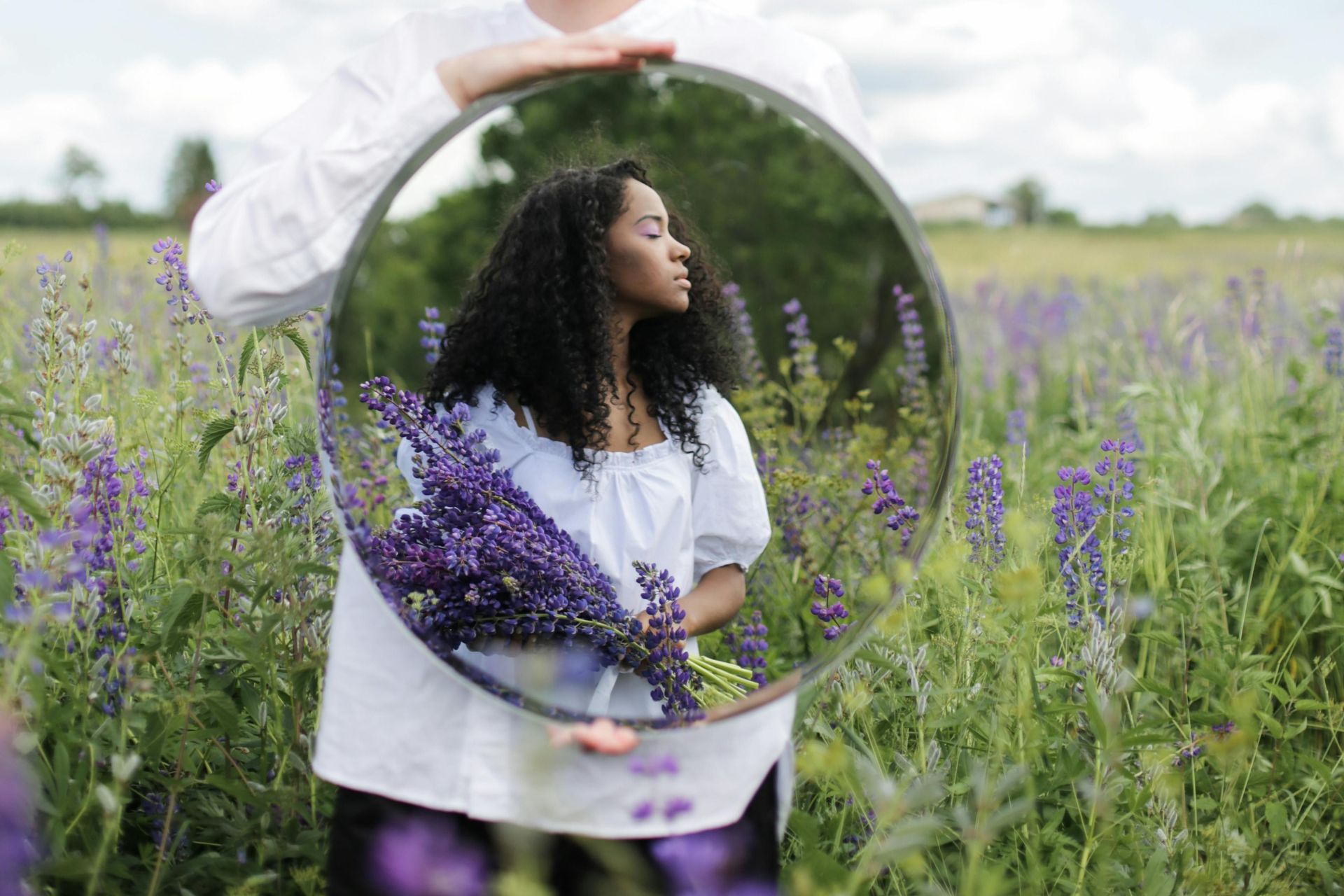 A Black woman standing in a field of lilacs, holding a mirror.