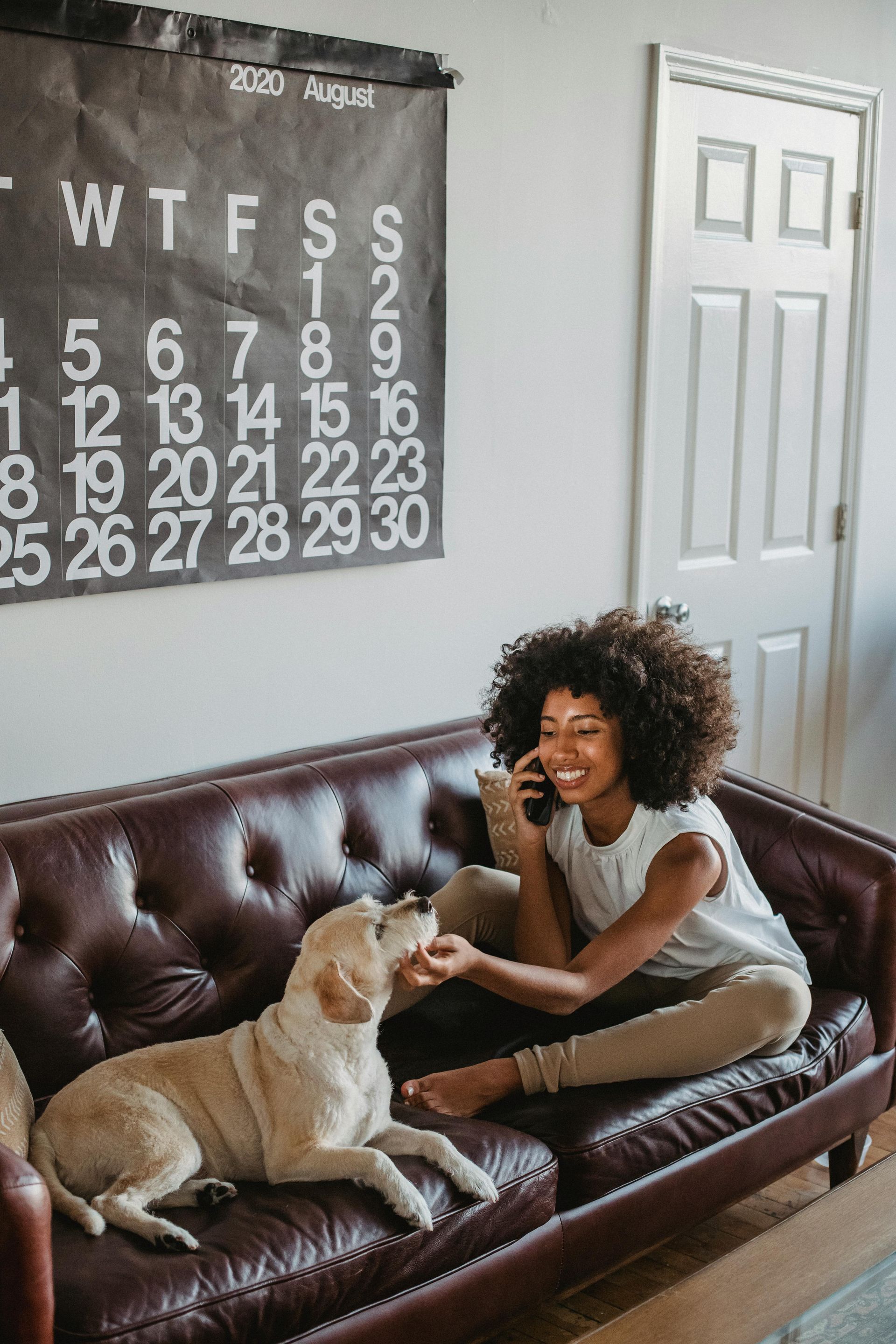 A woman is sitting on a couch with a dog and talking on a cell phone.