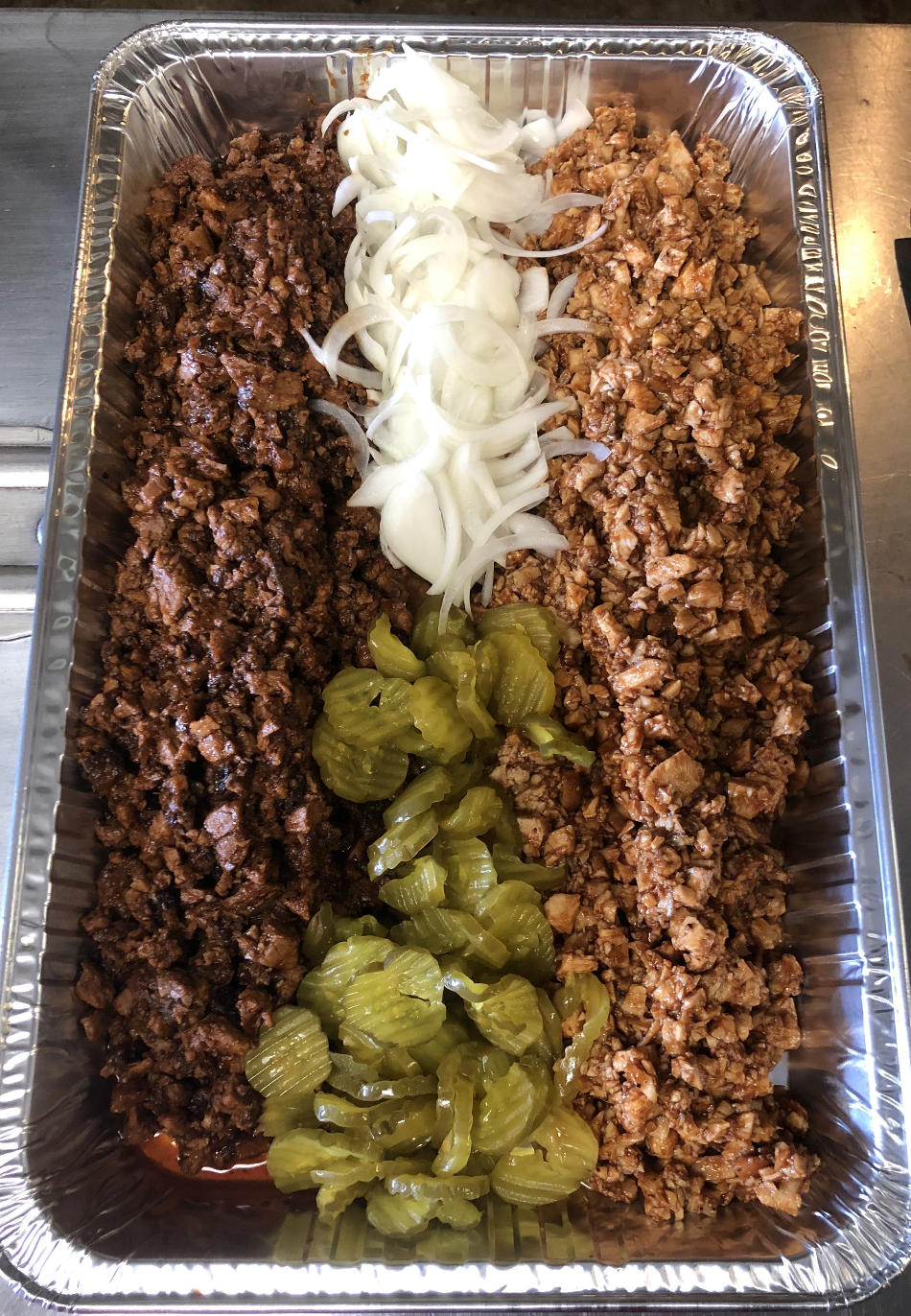 A tin foil tray filled with meat , pickles and onions.