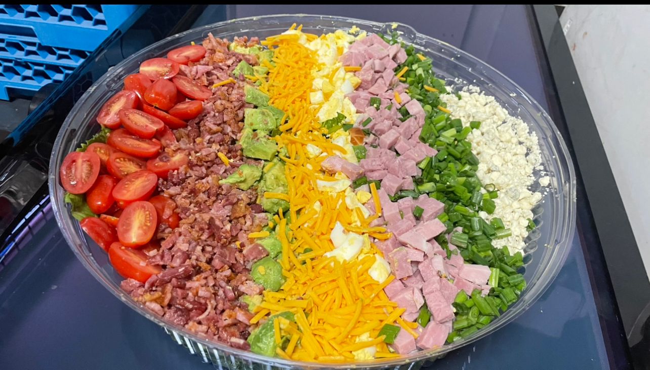 A salad in a plastic container with tomatoes , cheese , ham , and onions.