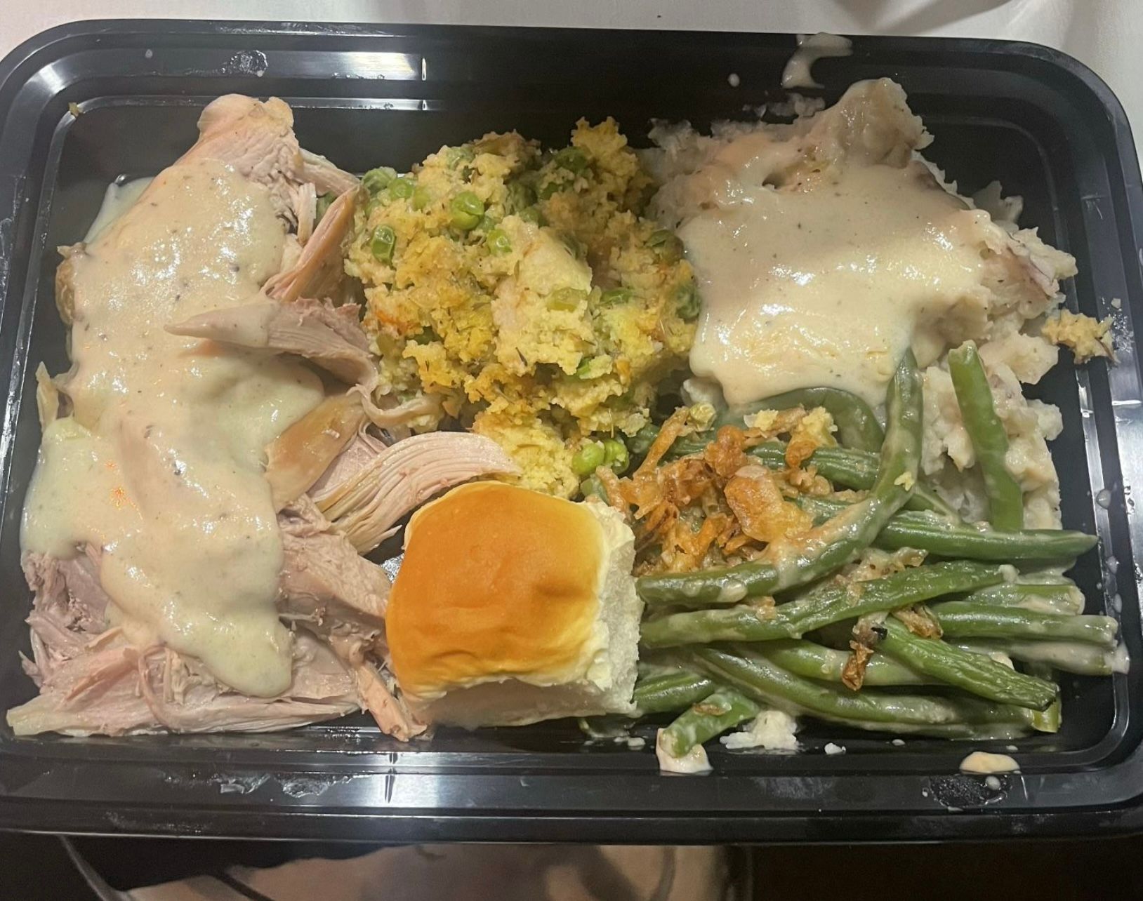 A plastic container filled with turkey , mashed potatoes , green beans and a bread roll.