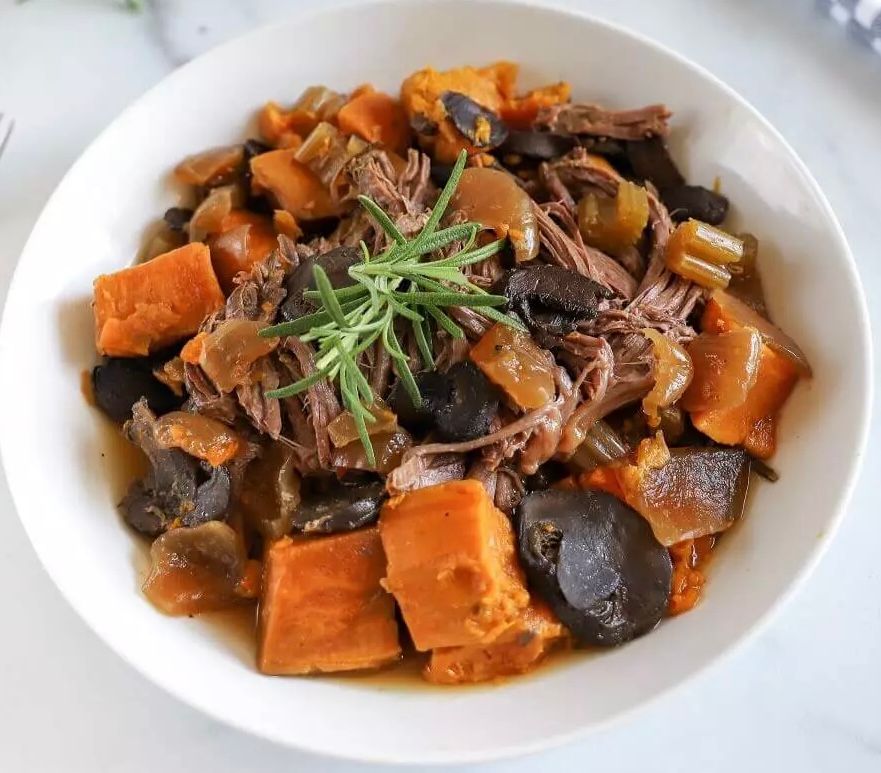 A bowl of beef stew with sweet potatoes and mushrooms on a table.