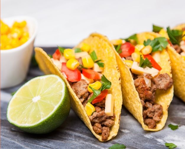 A row of tacos with meat , corn , tomatoes and limes on a table.