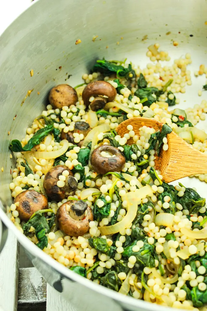 A pan filled with rice , mushrooms , spinach and onions with a wooden spoon.
