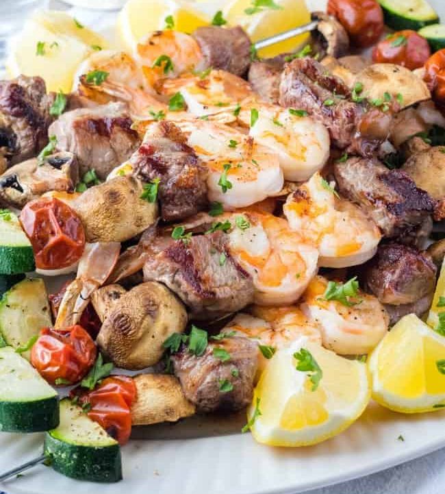 A white plate topped with meat , shrimp , vegetables and lemon slices.