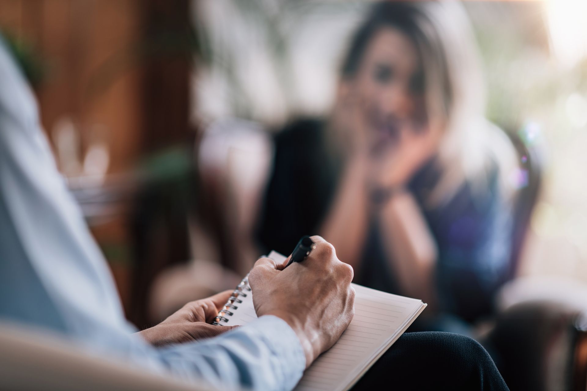Person writing in a notebook during a conversation in a blurred indoor setting