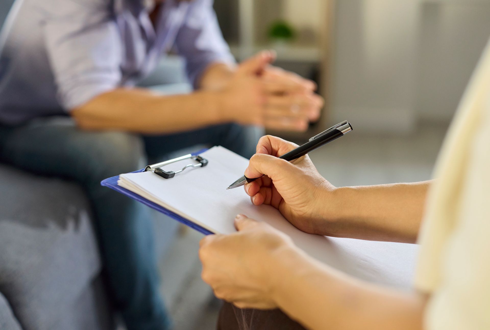 Therapist taking notes on a clipboard while meeting with a seated client in a 상담 room