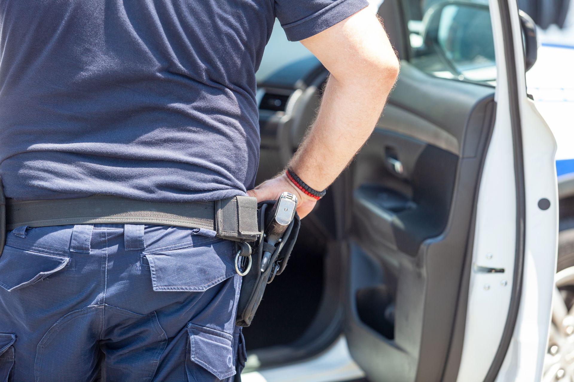 Police officer standing by an open car door, with a holstered gun visible at the waist.