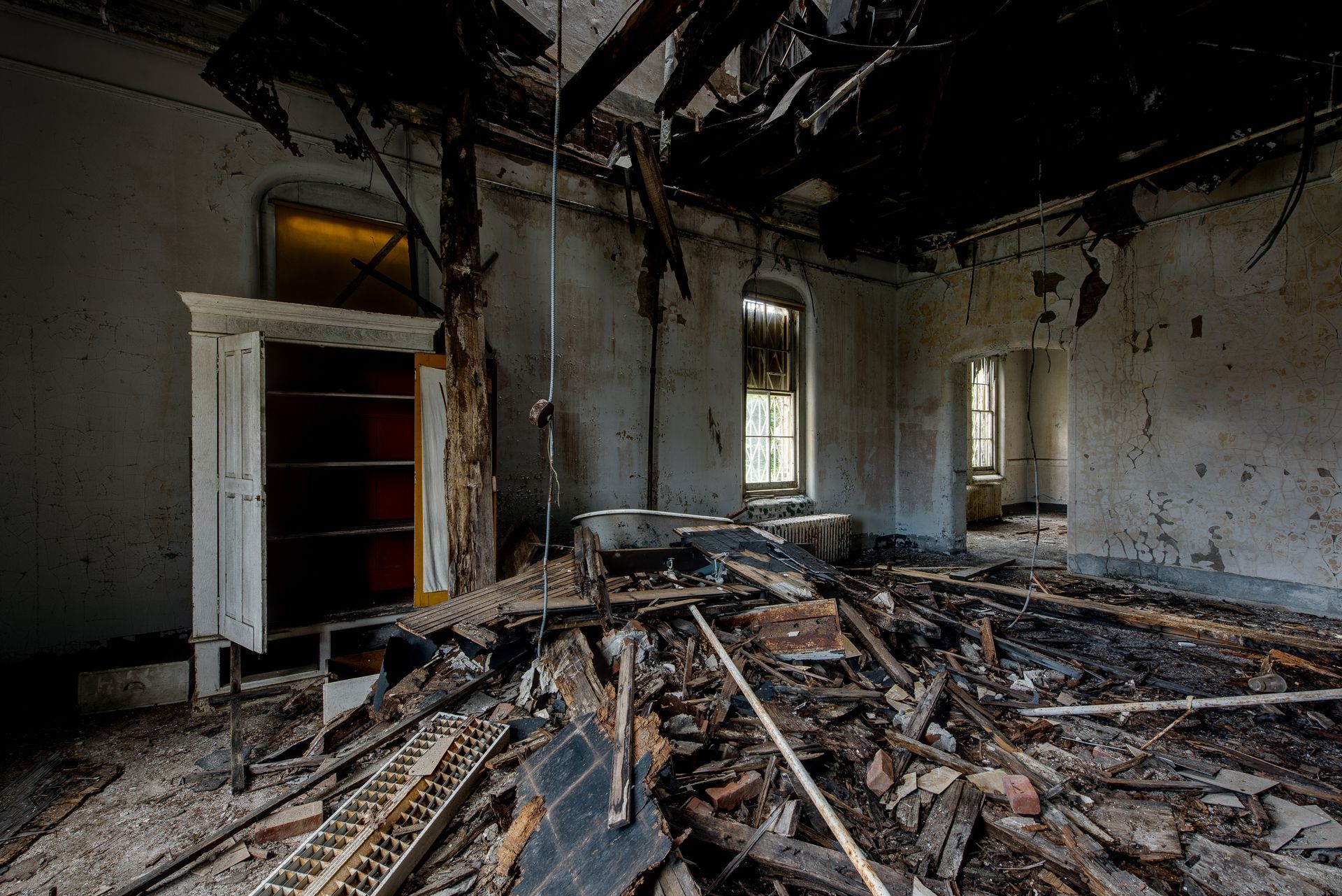 Interior view of a damaged room. Debris litters the floor, with a partially collapsed ceiling and an open white cabinet.
