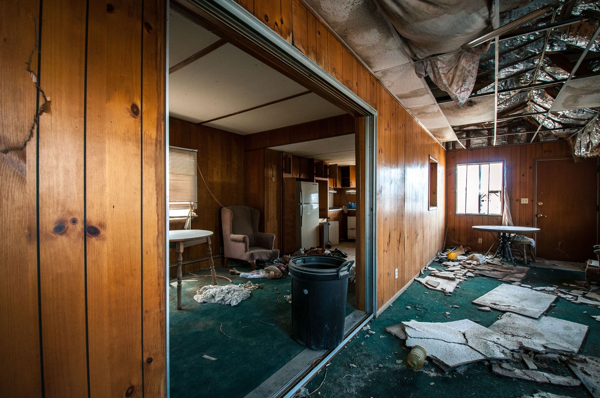 Interior of a dilapidated house, wood paneling, damaged ceiling, debris on the green carpet, doorway to a kitchen.