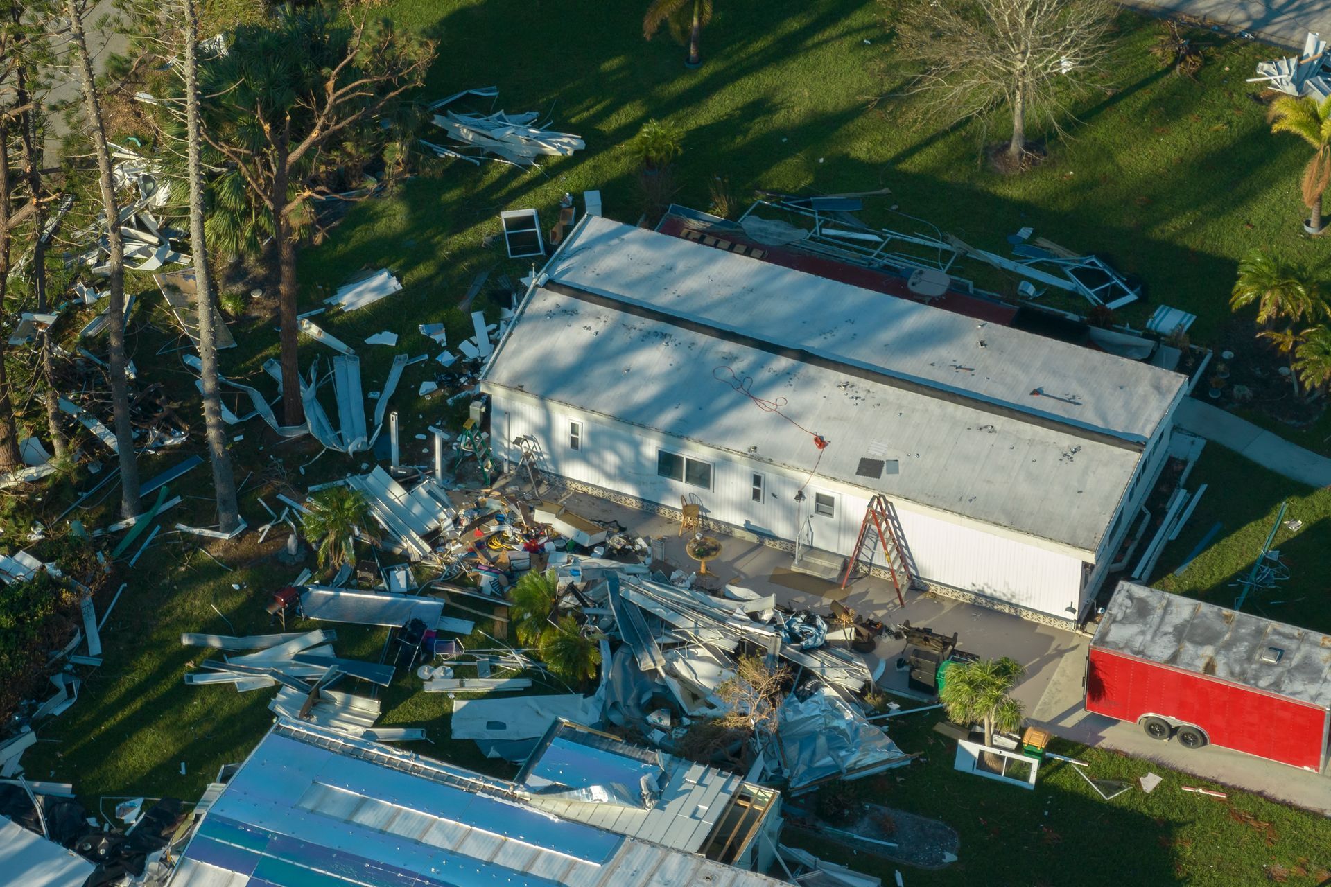 Aerial view of storm damage to a building, with debris scattered across a yard and a red trailer nearby