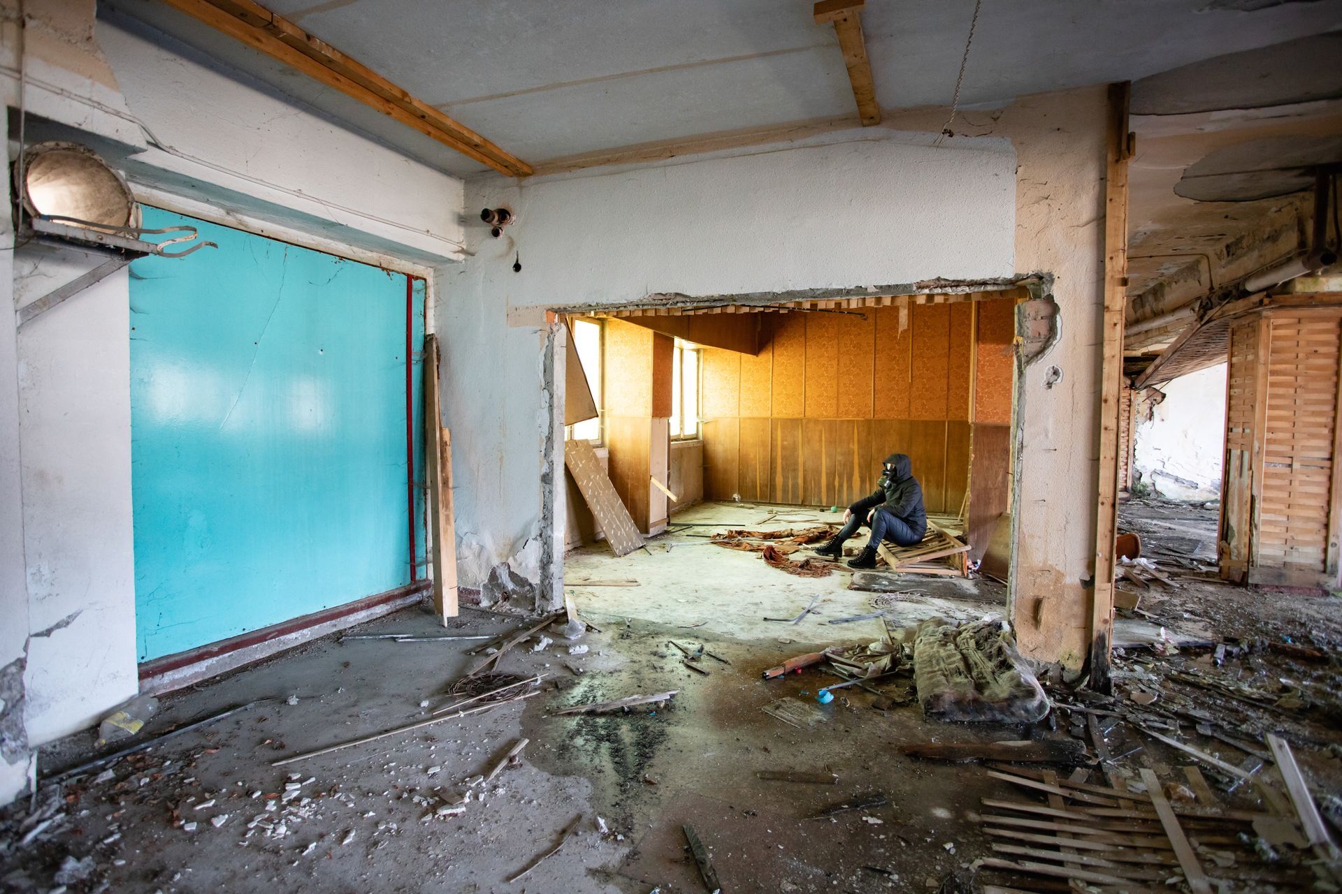 A person sits in a dilapidated room, blue door left. Debris litters the floor; walls are damaged.