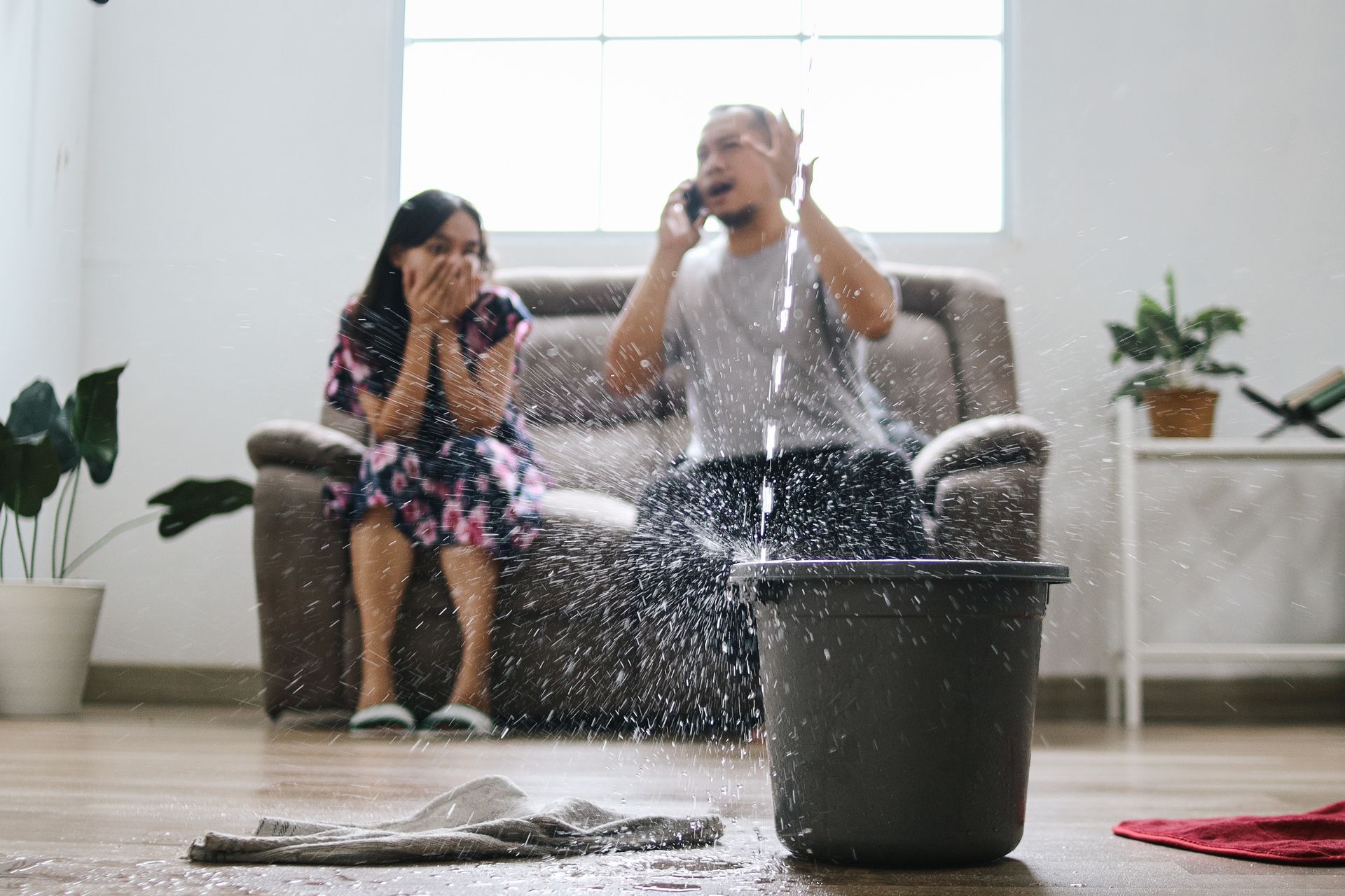 Couple reacting to water leaking on the floor, calling on phone while sitting on a couch. Bucket catching water.