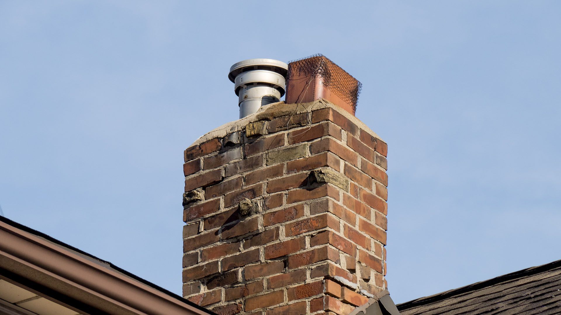Brick chimney with metal flue against a blue sky, mounted on a roof.