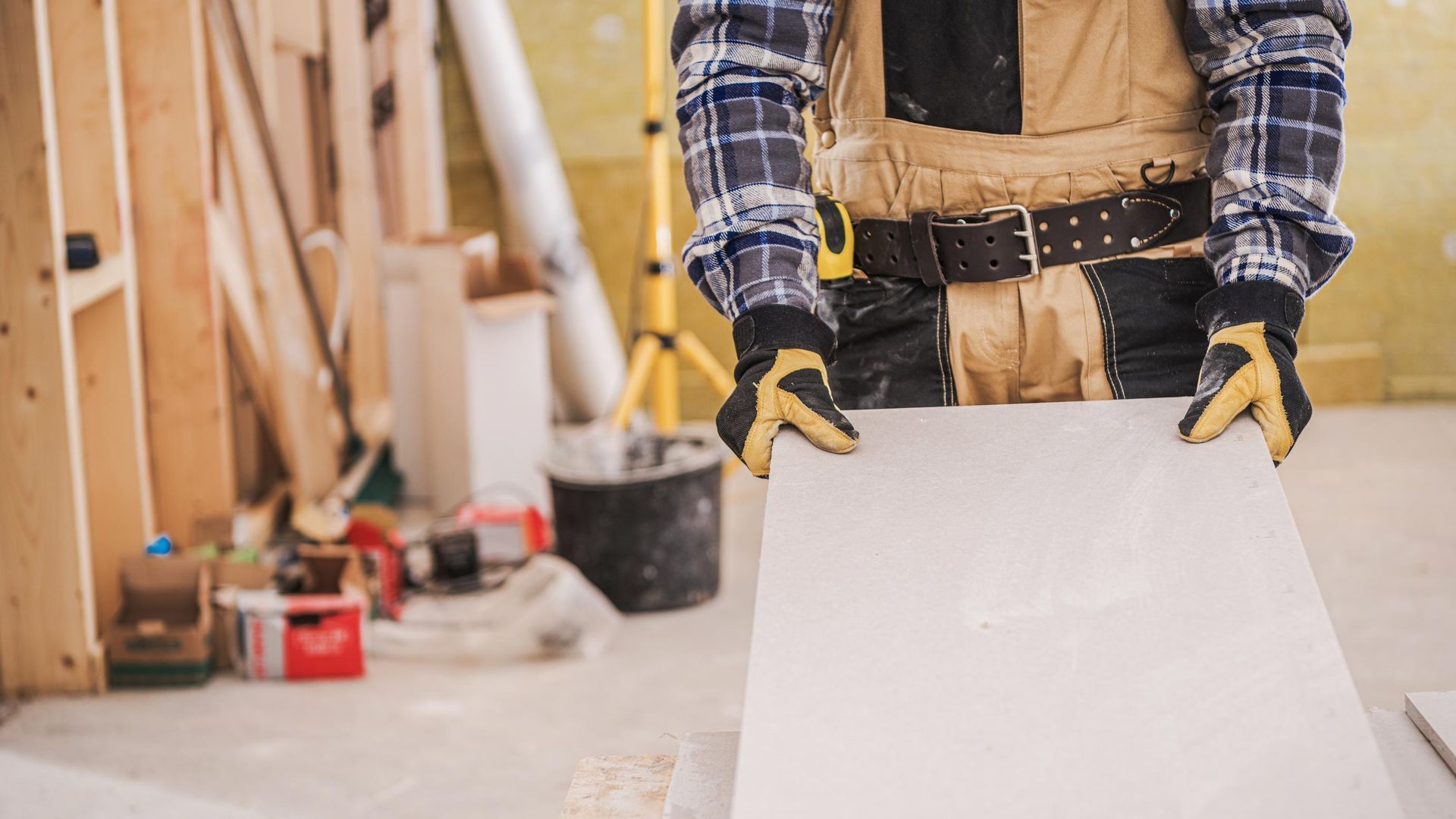A man is holding a piece of drywall in a room under construction.