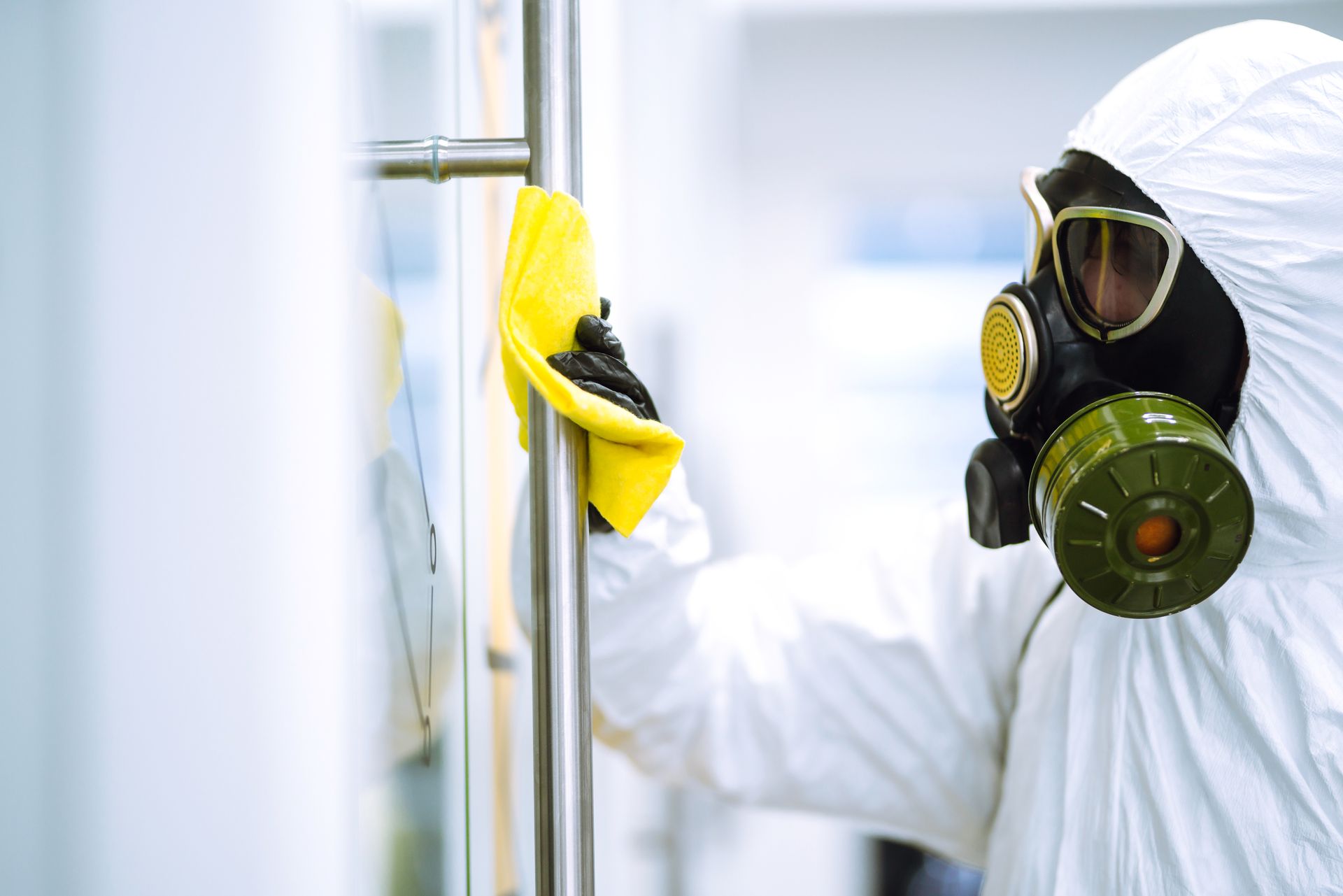 A person in a full white hazmat suit and gas mask wipes a metal door handle with a yellow cloth in a sterile setting.