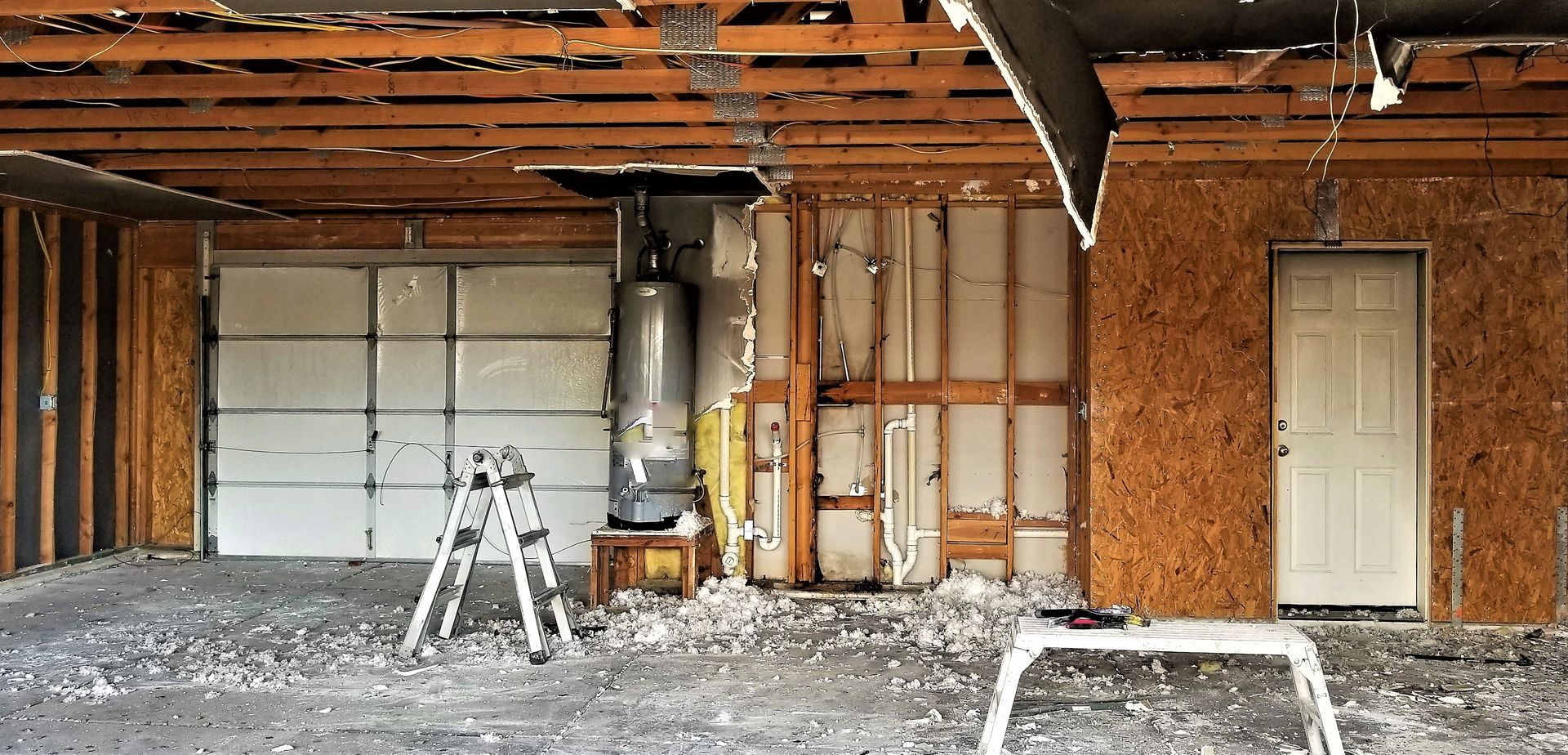 A garage undergoing demolition with exposed framing, a water heater, and a partially intact garage door and side door.