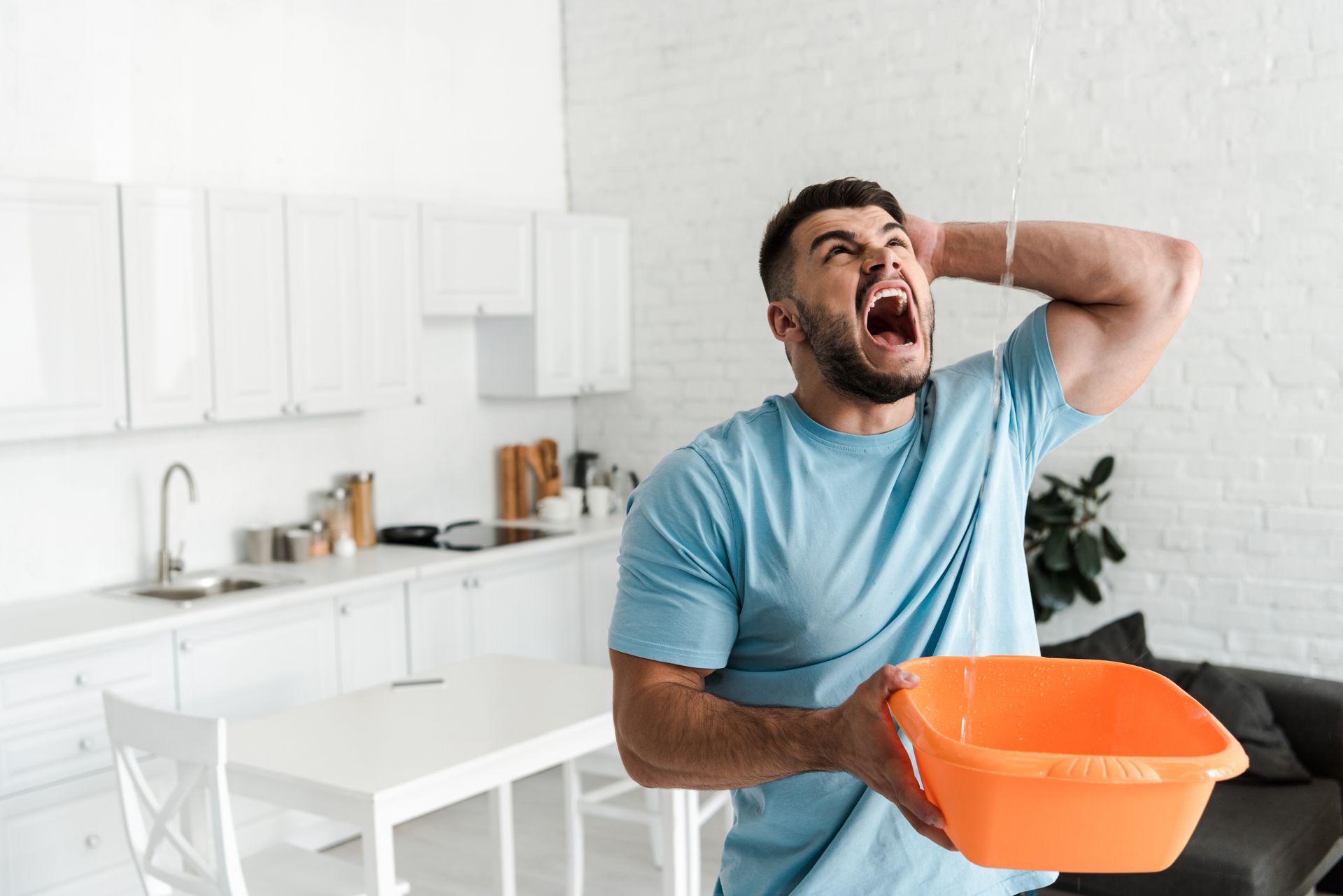 Man in blue shirt clutching his face and holding an orange basin in a bright kitchen