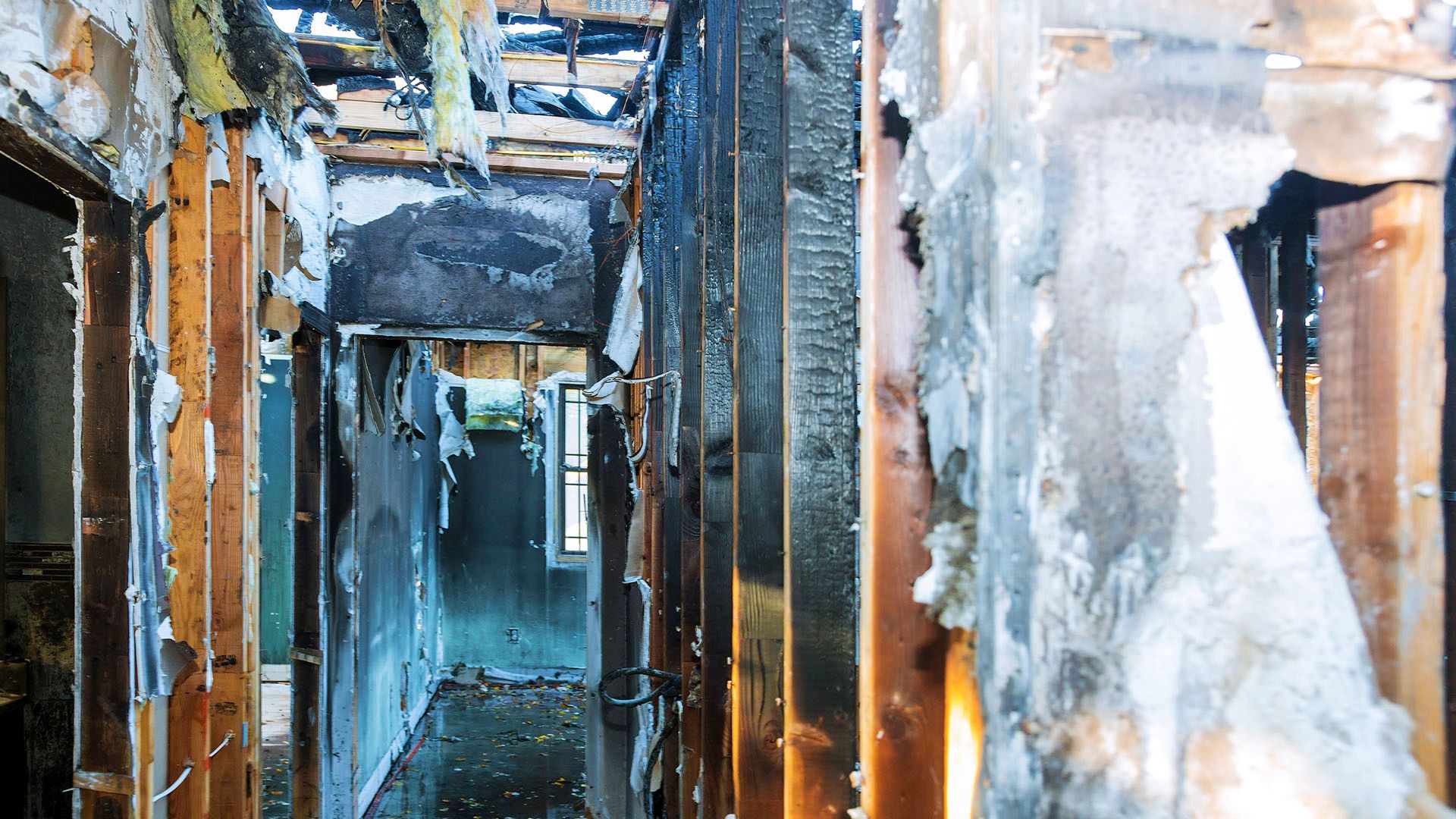 Hallway interior severely damaged by fire, with charred wood studs and debris.