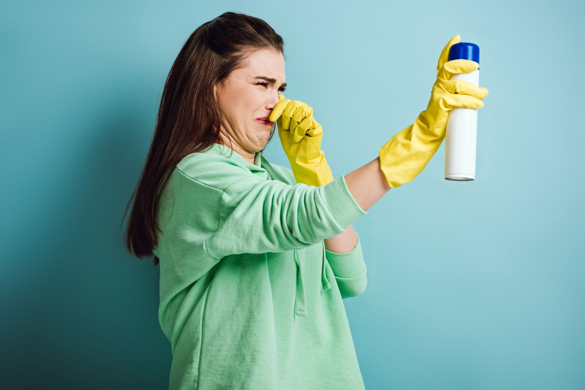 Woman in yellow gloves using a spray bottle and covering her nose against a blue background