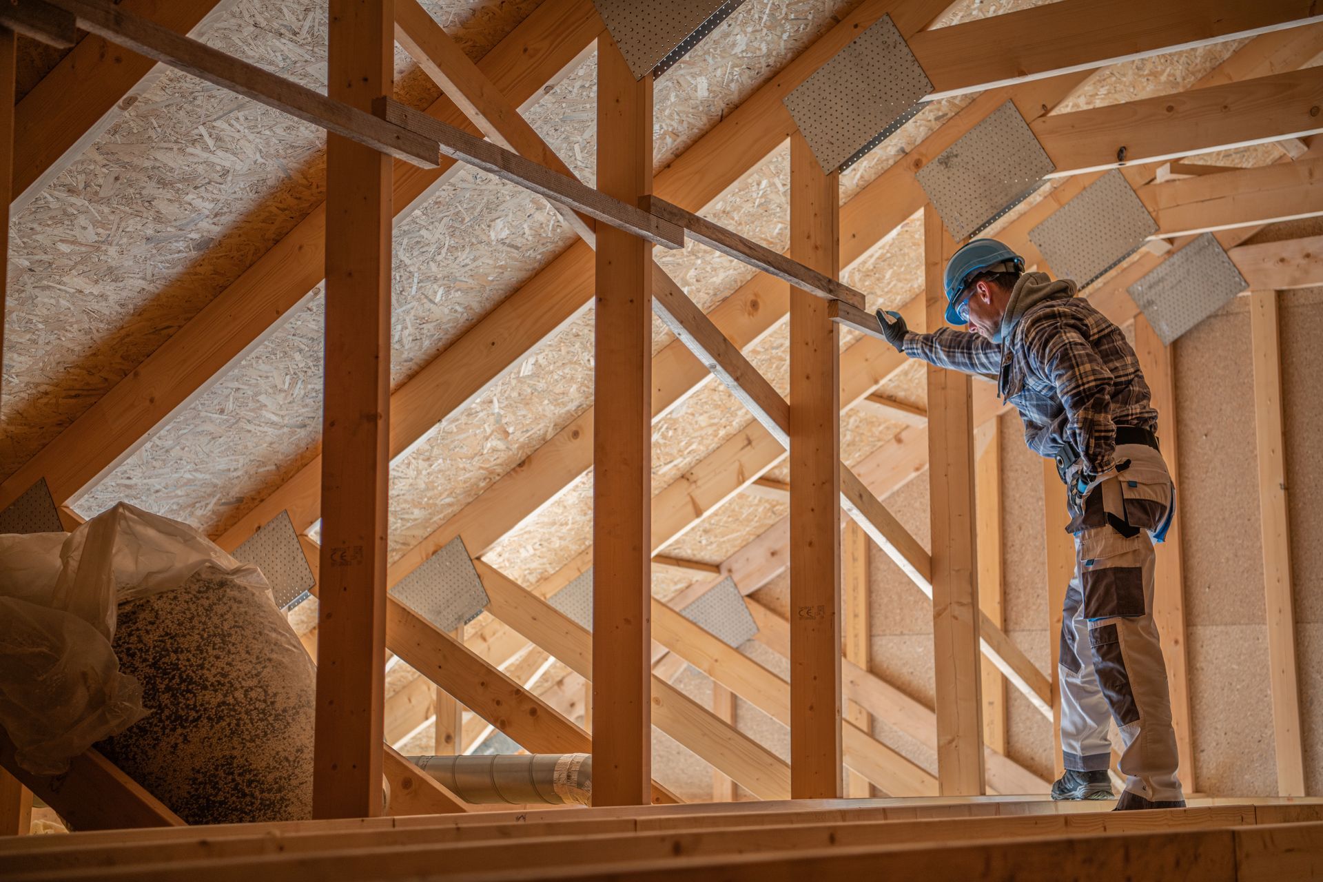 Man in a hard hat inspecting wooden attic framing. Bag of insulation in the background.