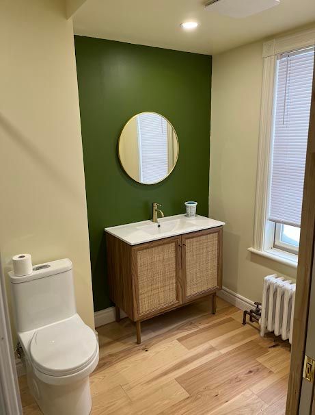 Une salle de bains avec un mur d'accent vert, un meuble-lavabo en bois et un miroir à cadre doré. Sol en bois clair.