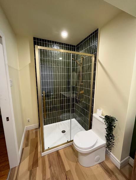 Salle de bains avec douche en verre à cadre doré, mur carrelé noir, toilettes blanches et sol imitation bois.