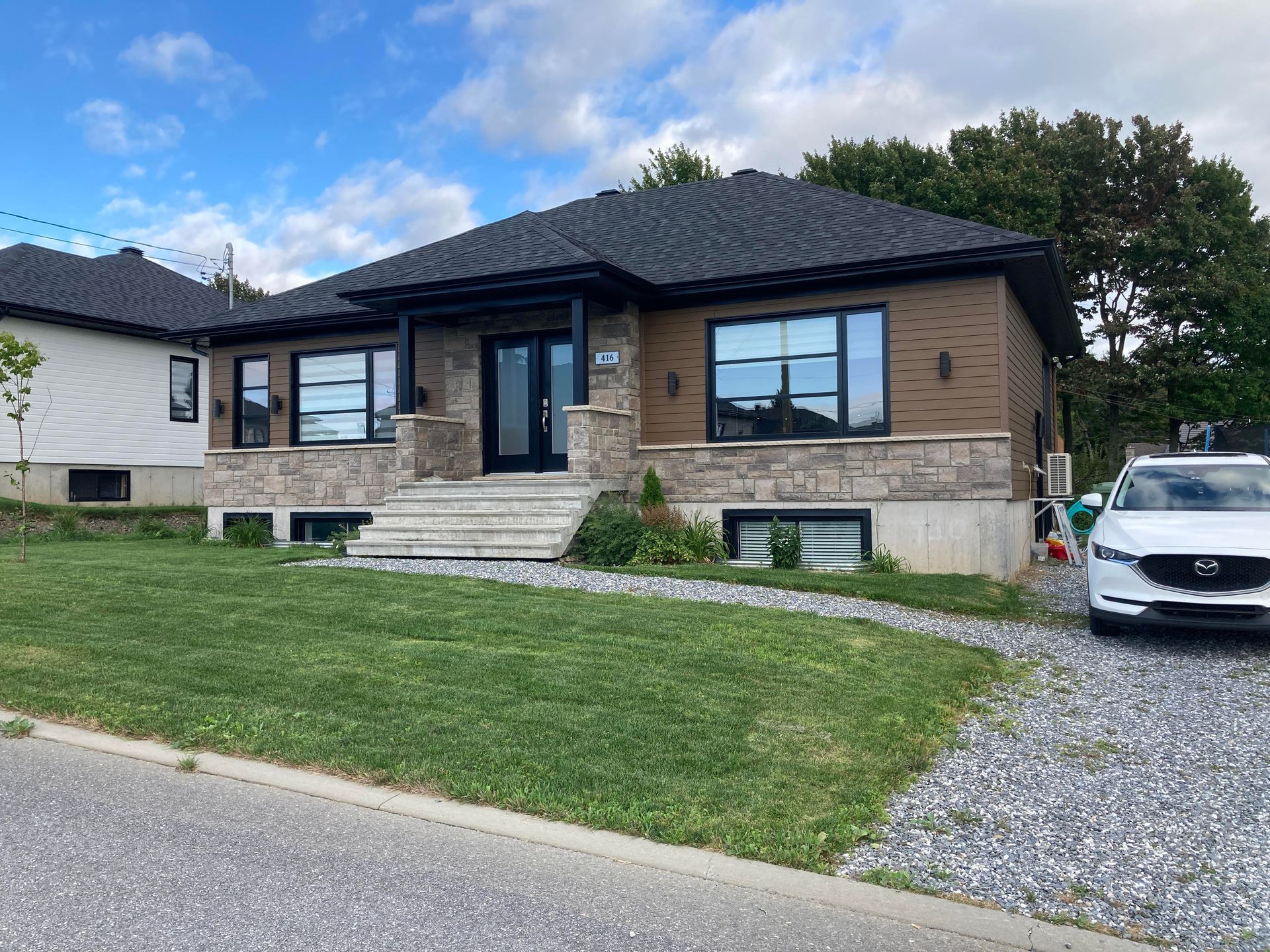 Maison en pierre avec bardage brun, toit sombre et pelouse verte. Une voiture est garée sur du gravier.