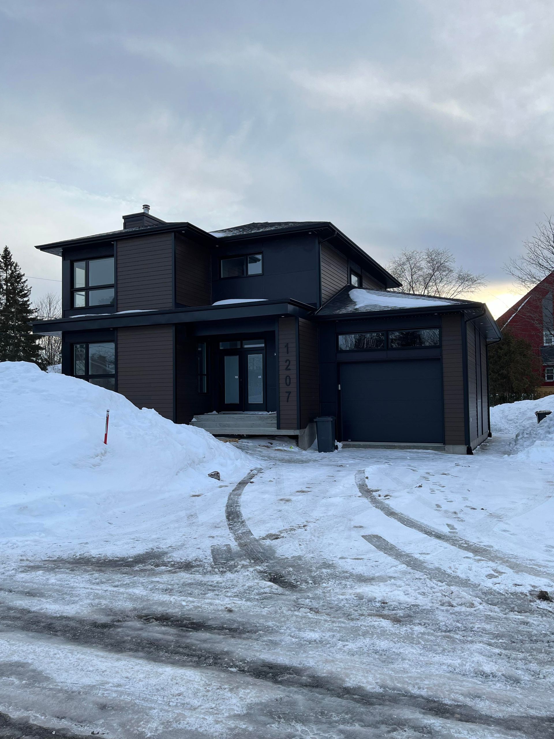 Maison moderne à deux étages avec bardage et garage foncés, allée enneigée, ciel couvert.