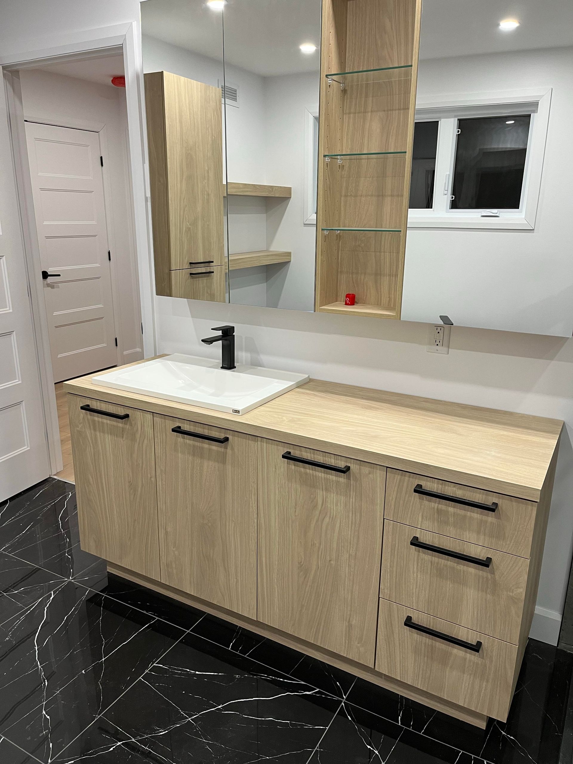 Salle de bains avec meuble-lavabo en bois clair, robinetterie noire et grand miroir.
