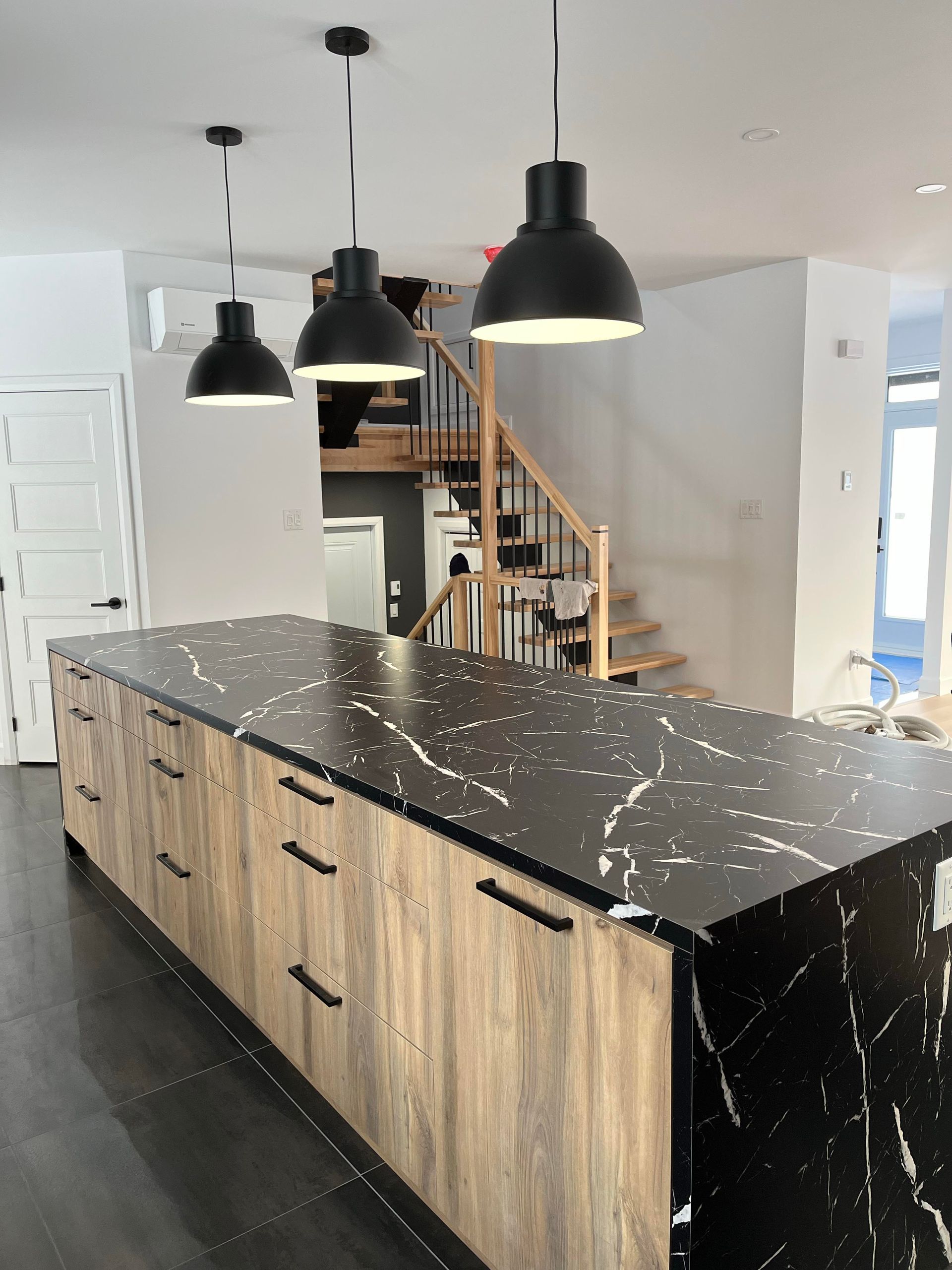 Îlot de cuisine avec comptoir noir, armoires couleur bois et suspensions noires. Escalier en arrière-plan.