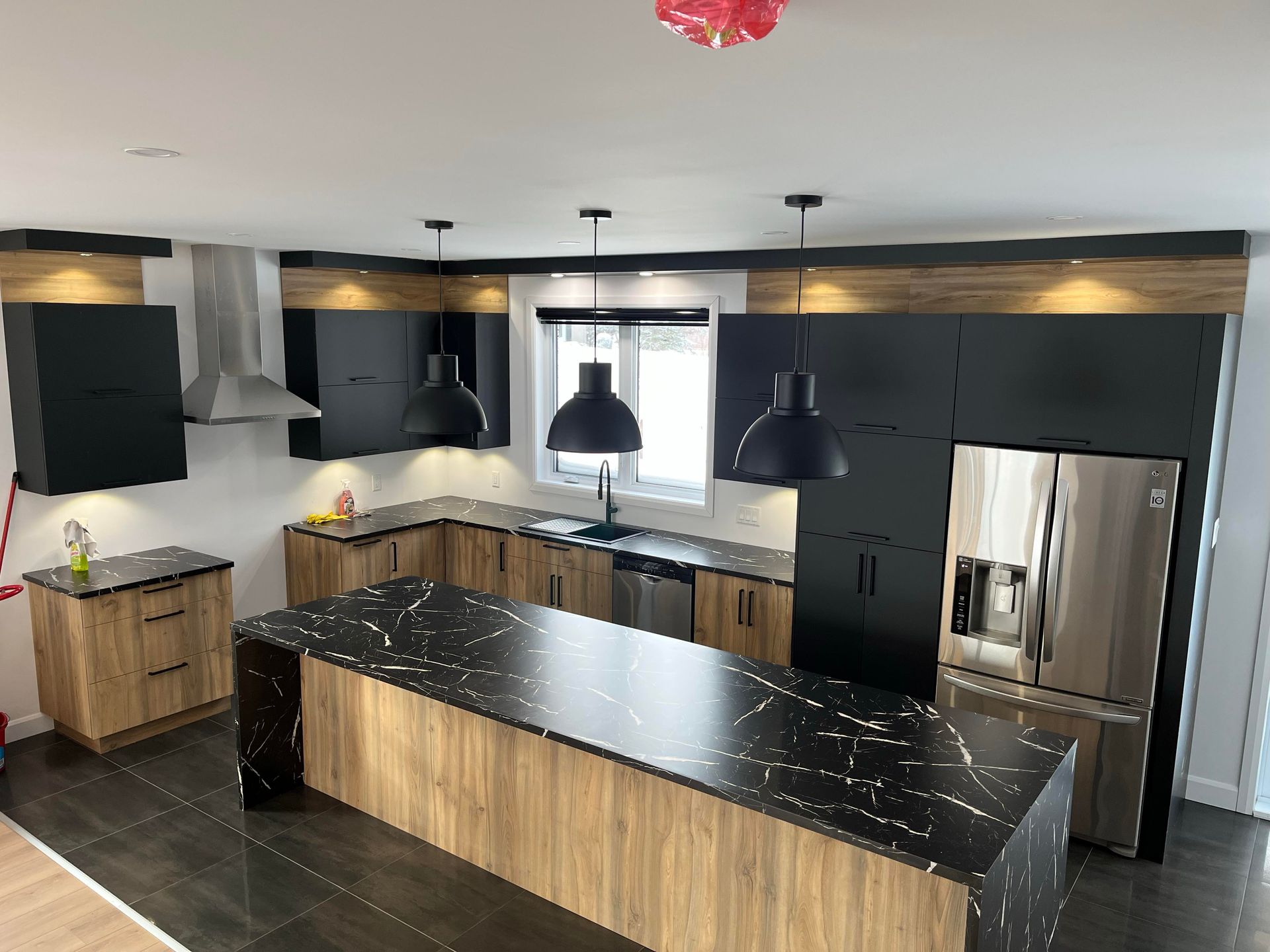 Cuisine moderne avec armoires noires et en bois, grand îlot avec comptoir noir, appareils électroménagers en acier inoxydable et luminaires suspendus.