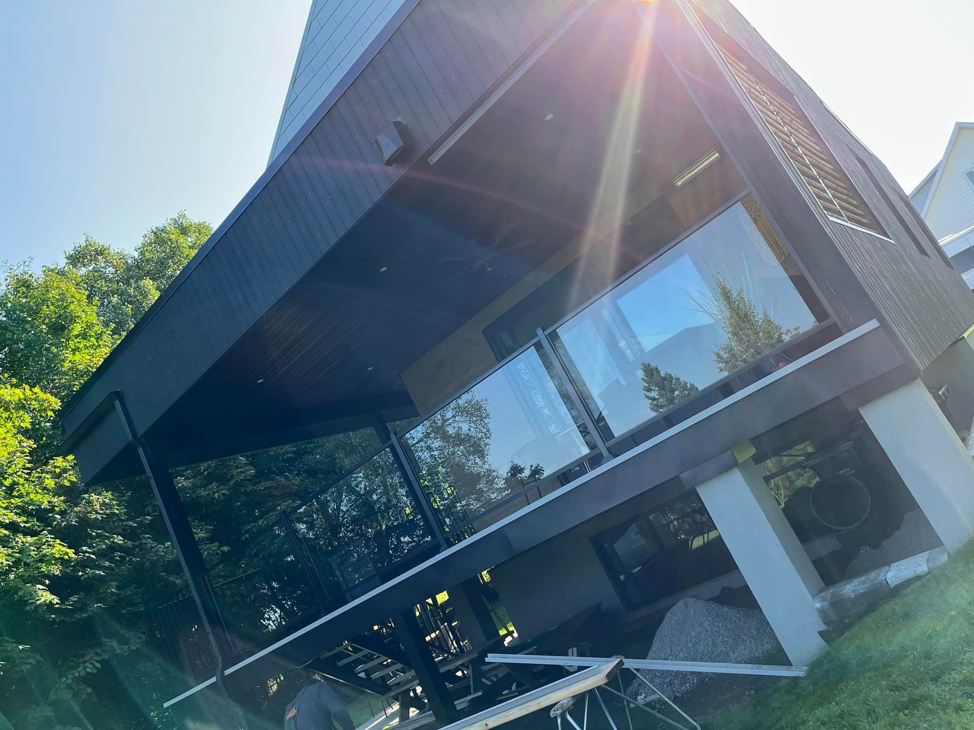 Maison moderne noire avec balustrades en verre, soleil éclatant et arbres.