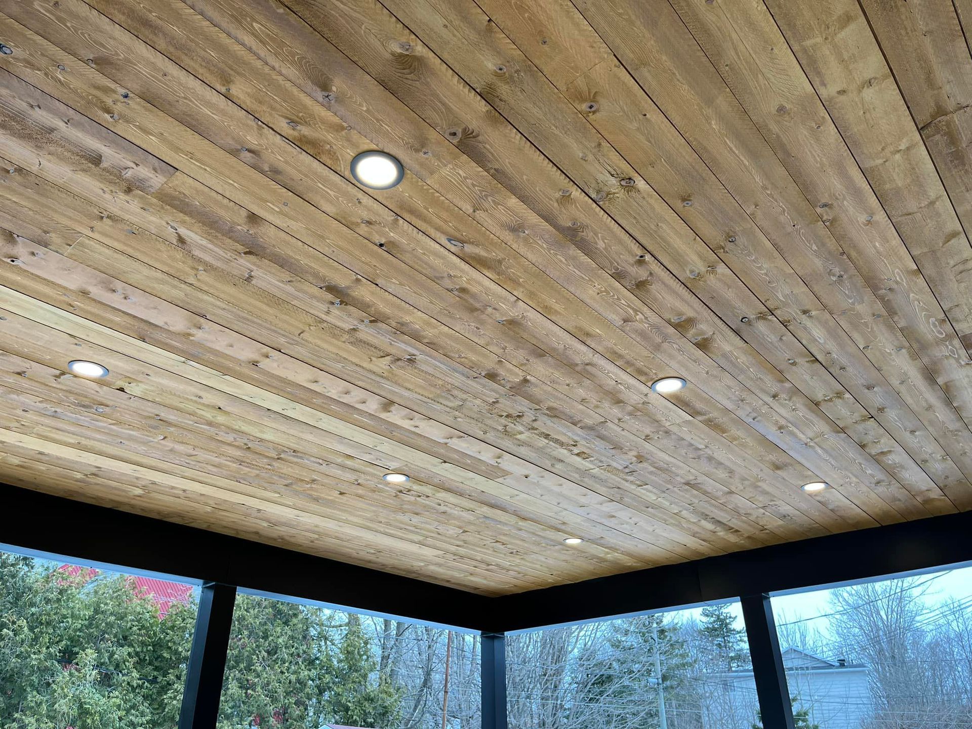Plafond en bois avec spots encastrés, vu d'en bas, au-dessus d'une véranda avec vue sur l'extérieur.