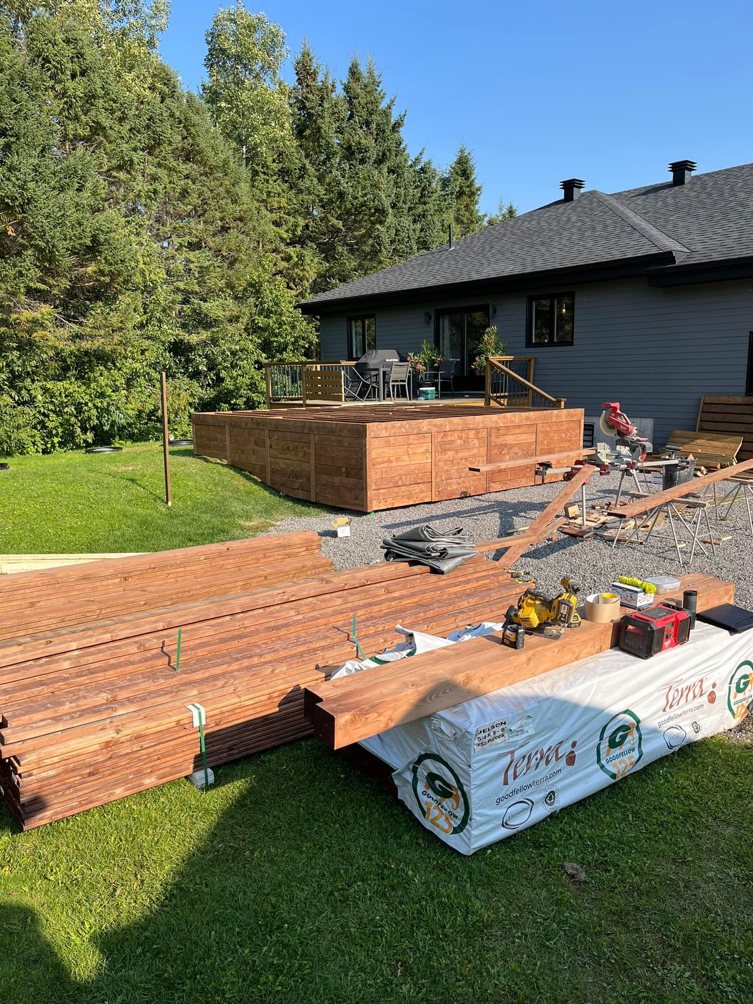 Construction d'une terrasse en bois à l'extérieur d'une maison ; des piles de bois et d'outils sont entreposées sur la pelouse.