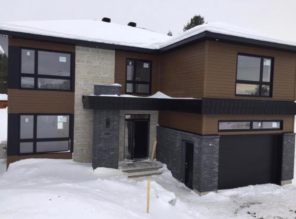 Maison moderne avec bardage brun, éléments en pierre, fenêtres noires et sol enneigé.