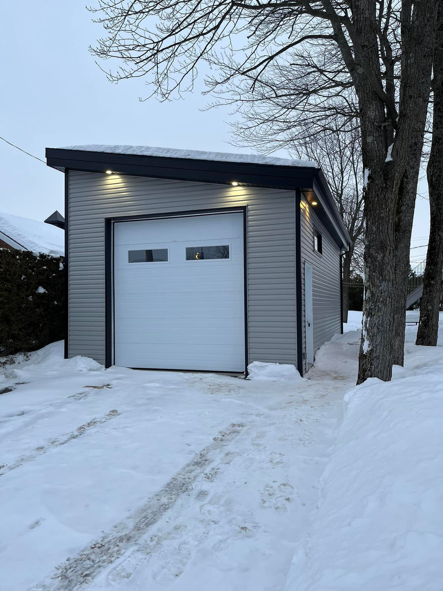 Garage avec porte blanche, allée enneigée, bardage gris et éclairage extérieur.