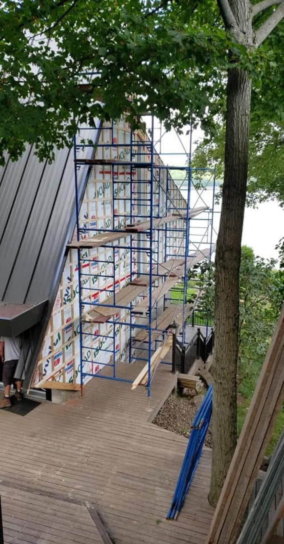 Échafaudages installés contre un bâtiment au toit noir, sur une terrasse en briques, près d'arbres. Travaux de construction en cours.