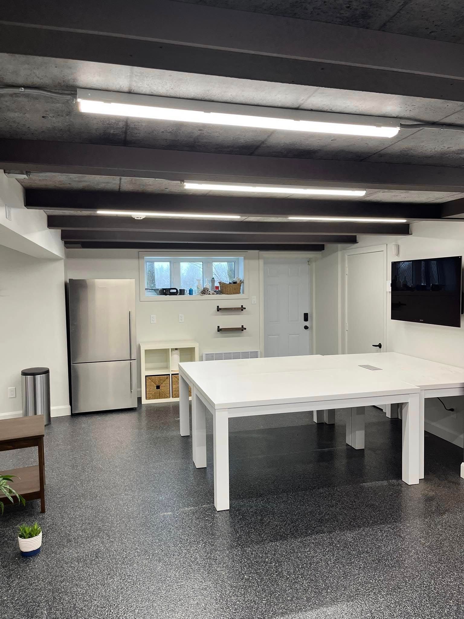 Pièce au sous-sol avec tables blanches, réfrigérateur, étagères, fenêtre et télévision. Poutres apparentes noires au plafond et sol moucheté.