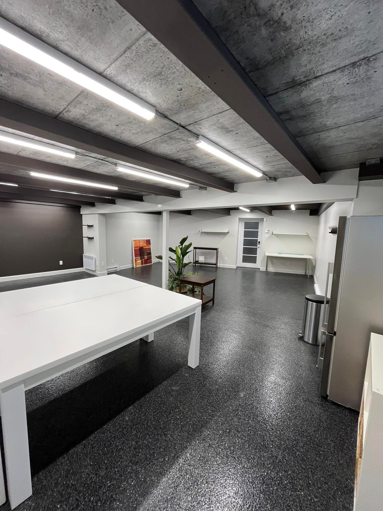 Vue intérieure d'un studio moderne avec des tables blanches, un sol sombre moucheté et un plafond en béton.