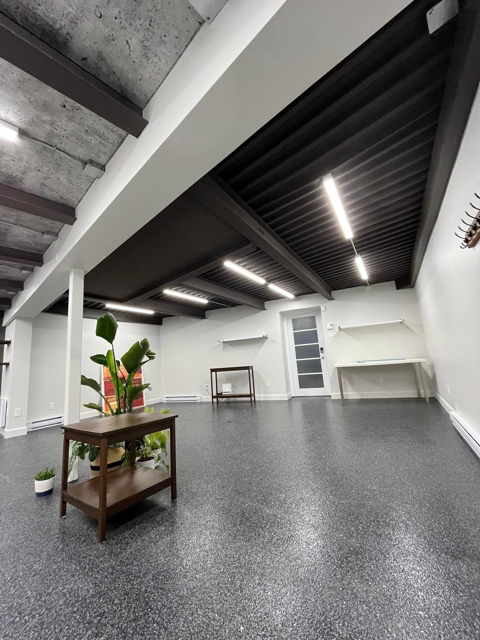 Pièce vide avec sol moucheté, murs blancs, plafond sombre et une table avec une plante.