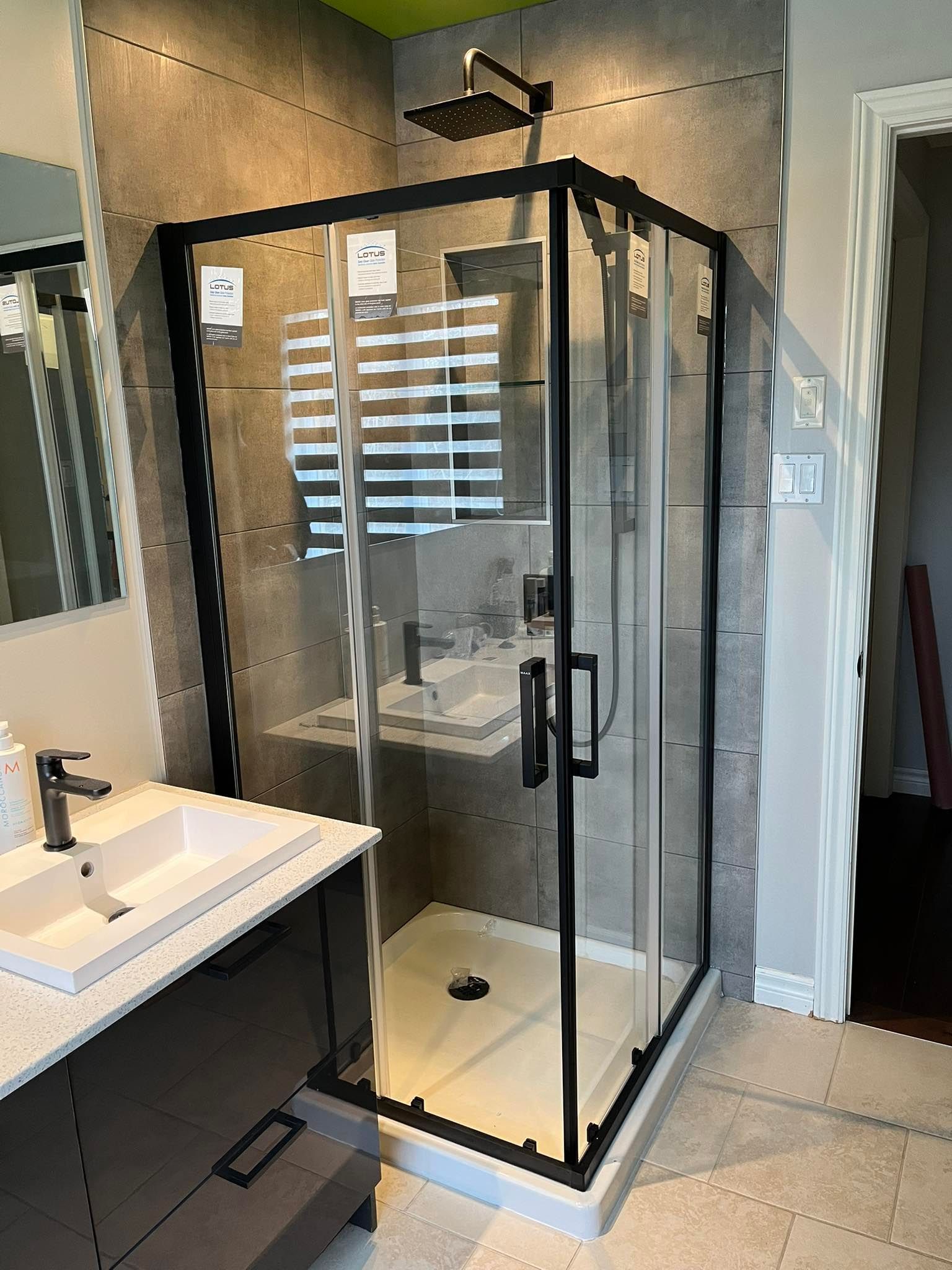 Salle de bains moderne avec douche en verre à cadre noir, mur carrelé gris, lavabo blanc sur meuble foncé et pommeau de douche carré à effet pluie.