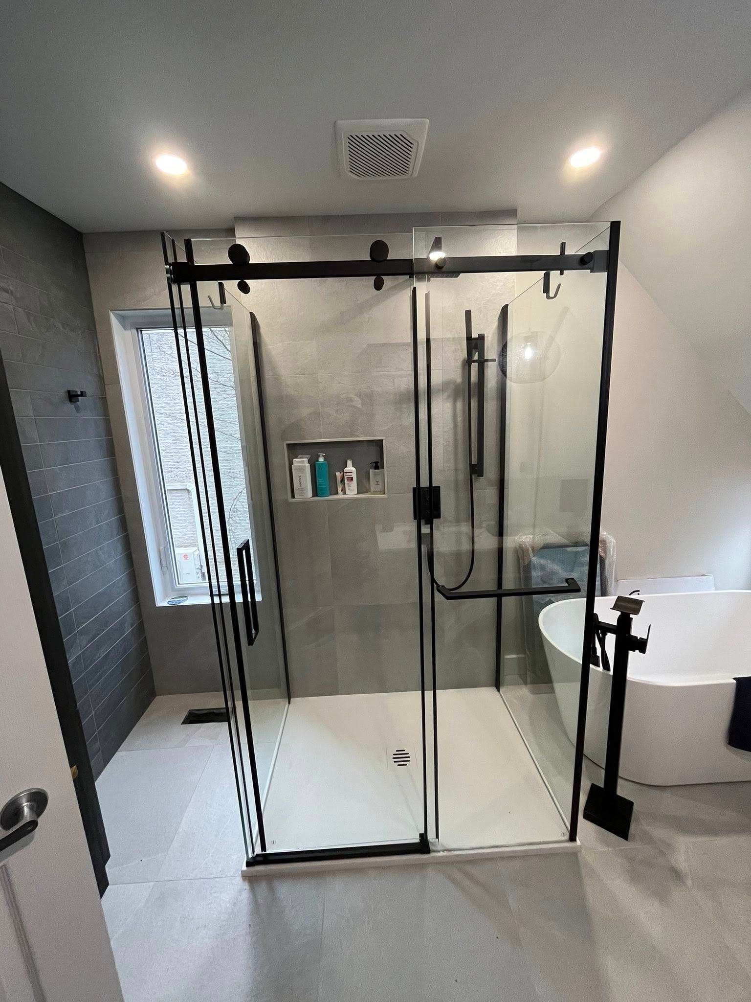 Salle de bains moderne avec une cabine de douche en verre, robinetterie noire, accessoires blancs et murs carrelés gris.