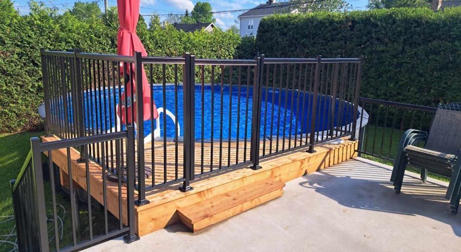 Piscine hors sol sur une terrasse en bois entourée d'une clôture noire et de verdure.