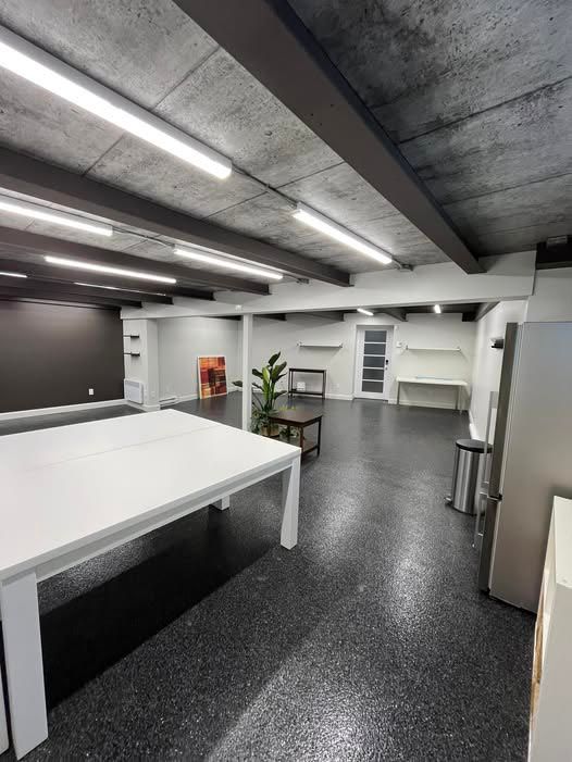 Espace de bureaux moderne, sol gris foncé, tables blanches, éclairage fluorescent, poutres apparentes et un mur noir.