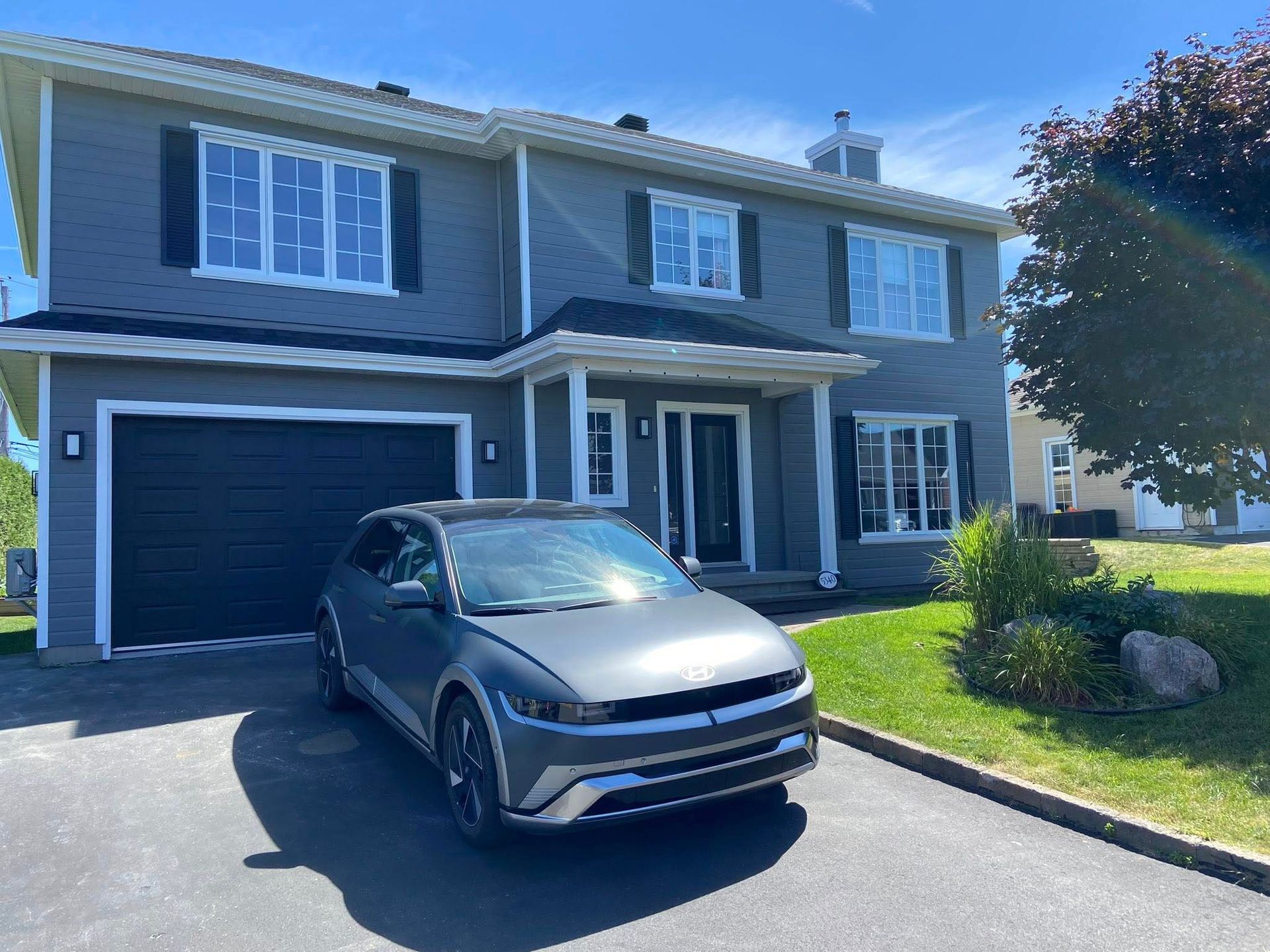 Maison grise avec volets noirs et garage, une voiture grise garée devant par une journée ensoleillée.