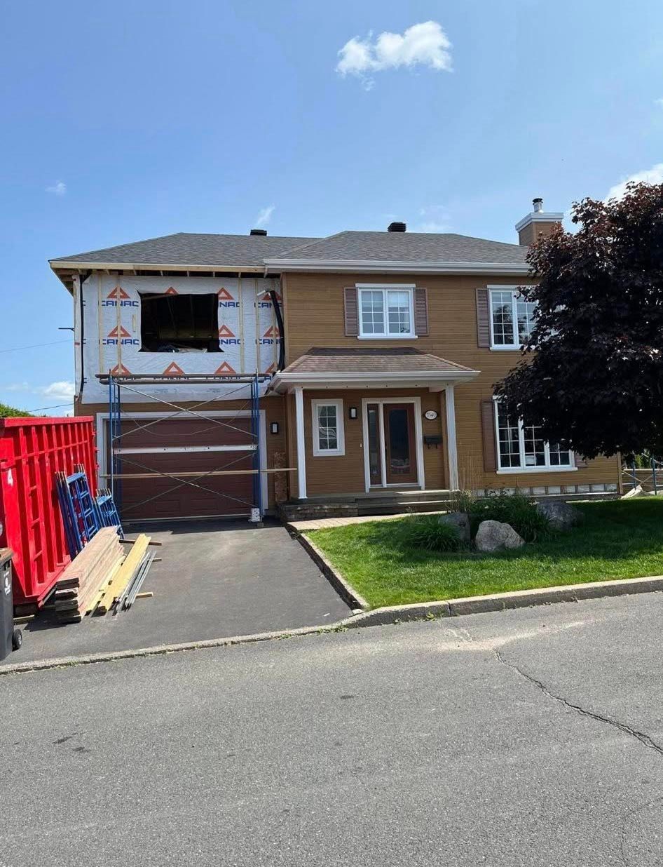 Maison en rénovation ; la charpente en bois est visible dans une partie inachevée. Bardage brun, porte de garage, ciel bleu.