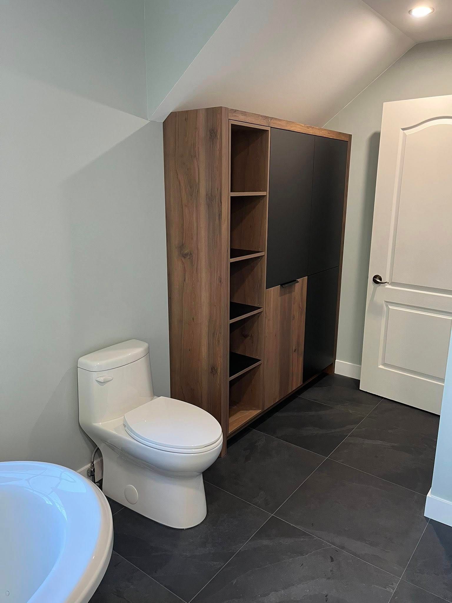Salle de bains avec toilettes, carrelage foncé au sol, armoire de rangement marron et porte blanche.