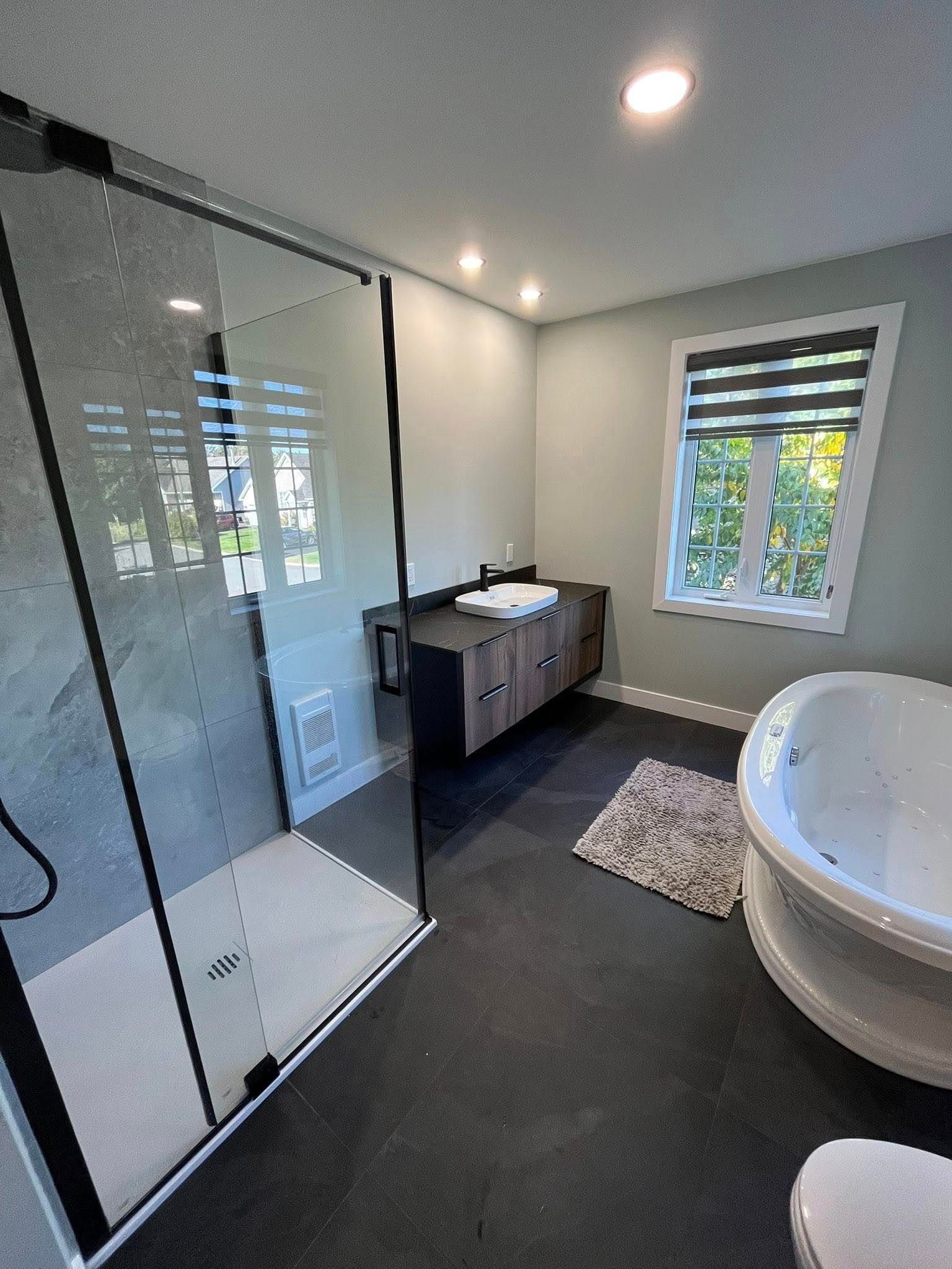 Salle de bains moderne avec douche en verre, sol foncé, meuble-lavabo et baignoire ronde près d'une fenêtre.