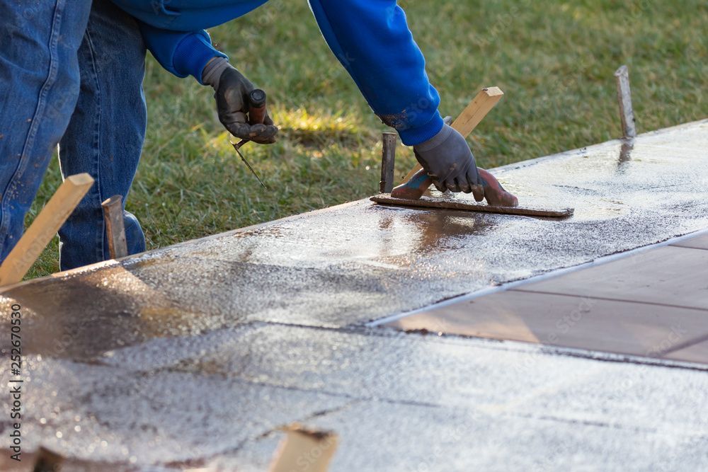 Une personne lisse du béton frais à la truelle à l'extérieur, des guides en bois sont visibles.